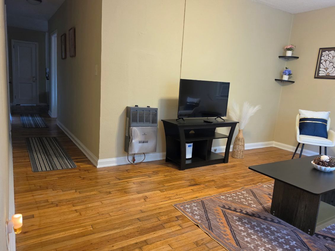 Una sala de estar con televisión sobre un mueble bajo, una mesa de comedor con un tazón, una silla y un tapete estampado.