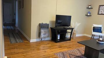 Una sala de estar con televisión sobre un mueble bajo, una mesa de comedor con un tazón, una silla y un tapete estampado.