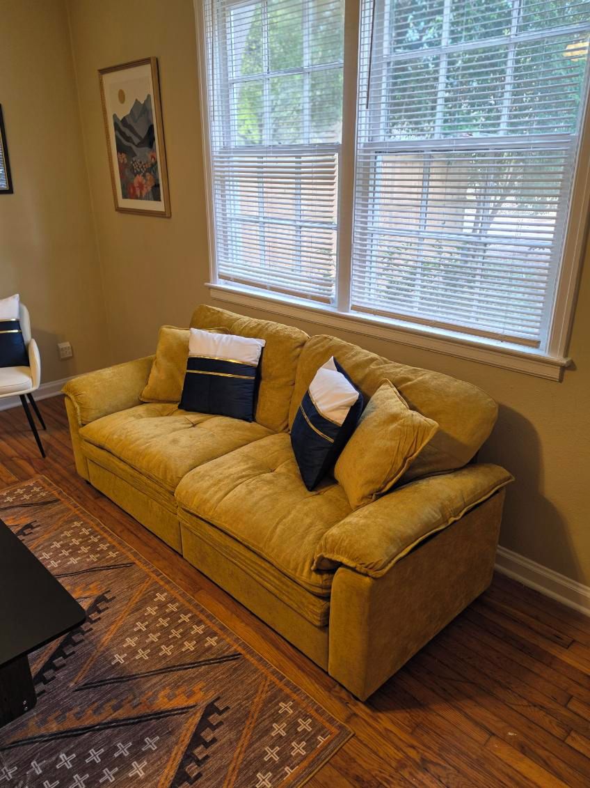Una habitación con sofá amarillo, alfombra estampada y ventana con persianas.