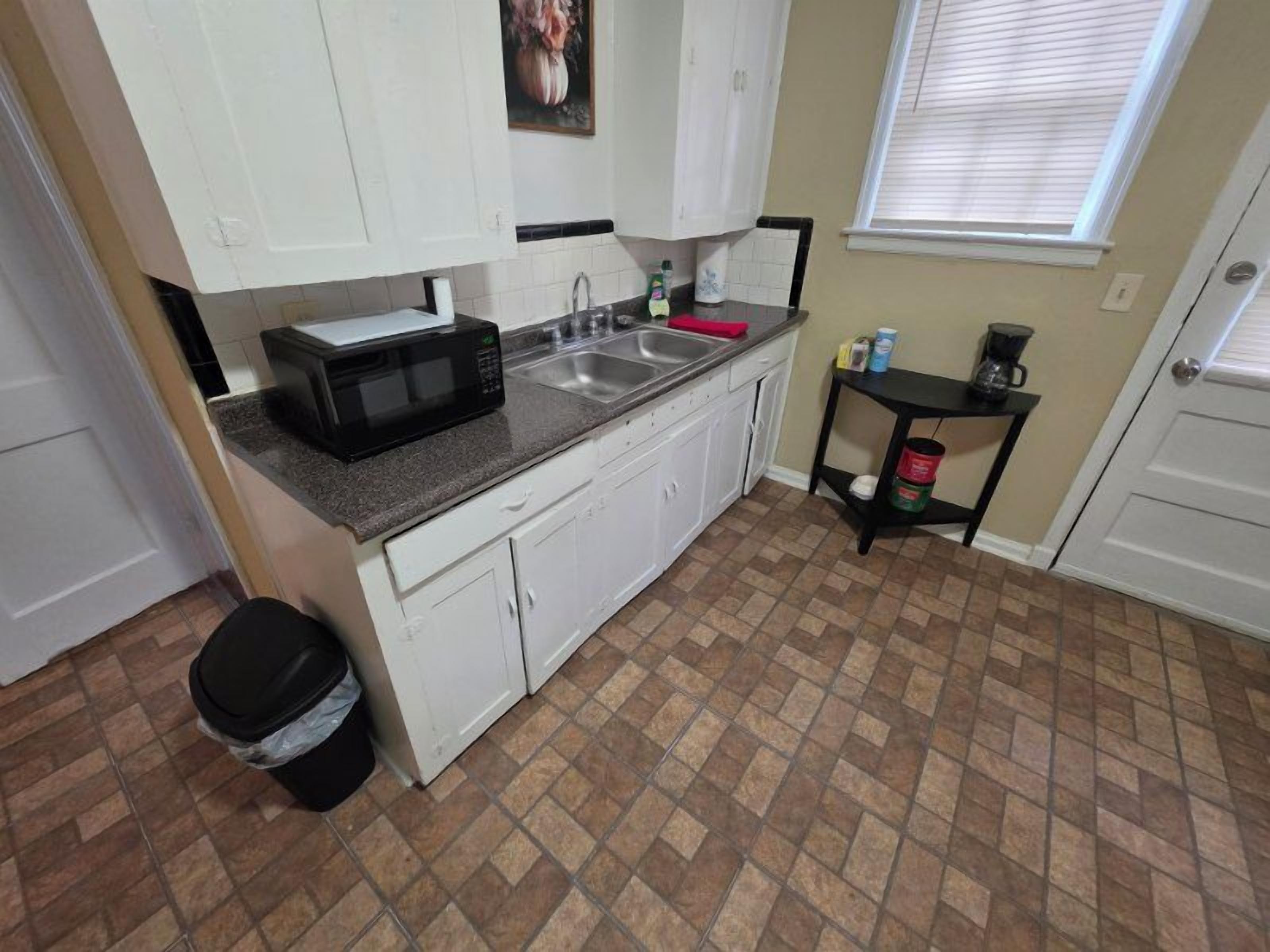 A compact kitchen with white cabinets, a granite countertop, a microwave, a sink, and a small black table with a coffee maker.
