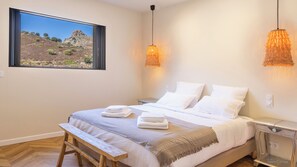 Quarto com uma cama grande, um banco, uma mesa de cabeceira, um criado-mudo, uma janela com vista para a montanha e duas luminárias pendentes.