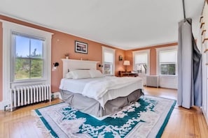 A bedroom with a bed, a radiator, a window, a lamp, and a view of greenery outside. - Port Clyde Road (Tenants Harbor)