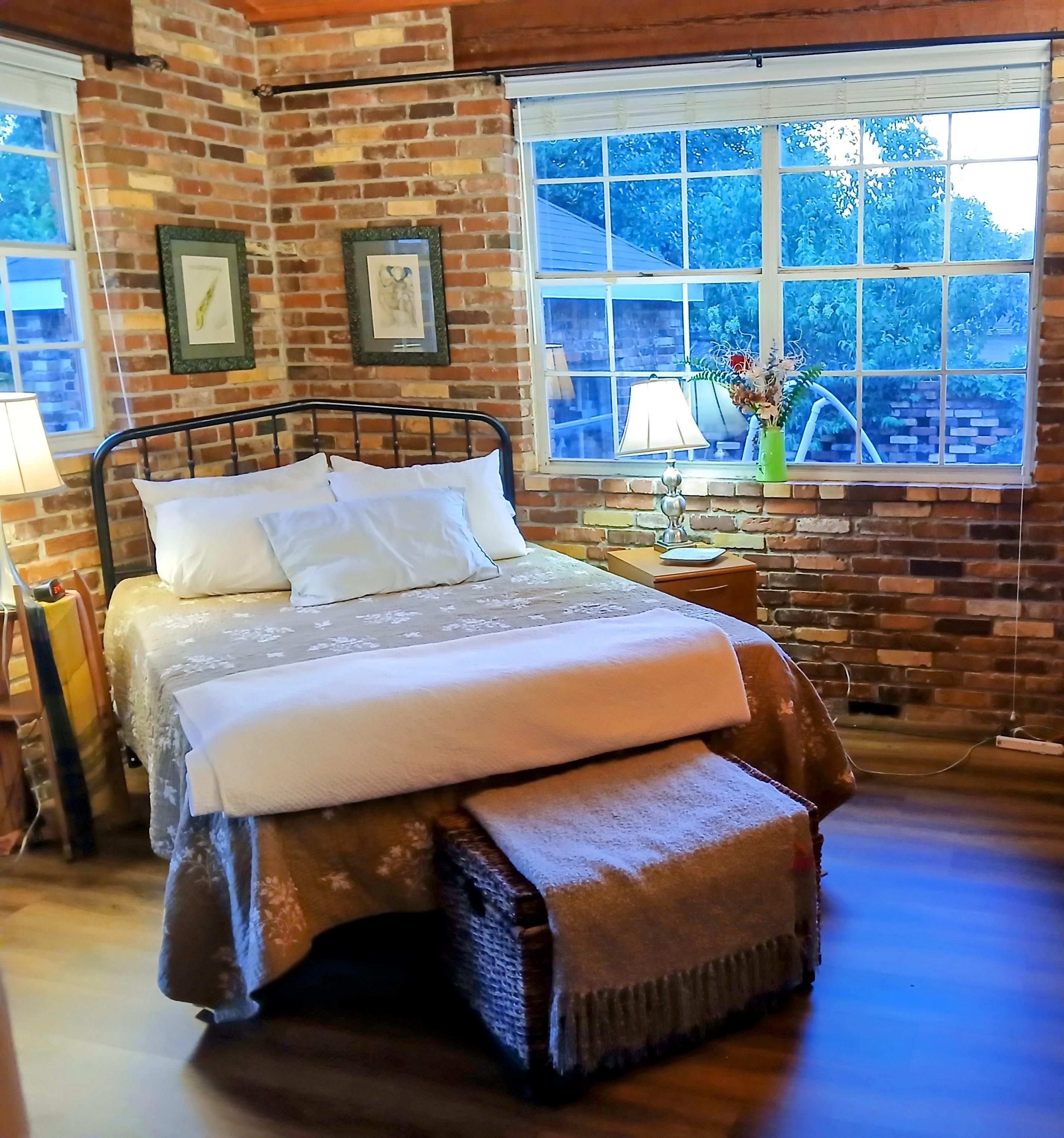 A room with a brick wall, a bed with white bedding, a window with a view of trees, and a nightstand with a lamp.