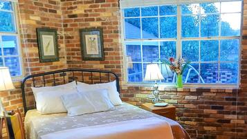 A room with a brick wall, a bed with white bedding, a window with a view of trees, and a nightstand with a lamp.