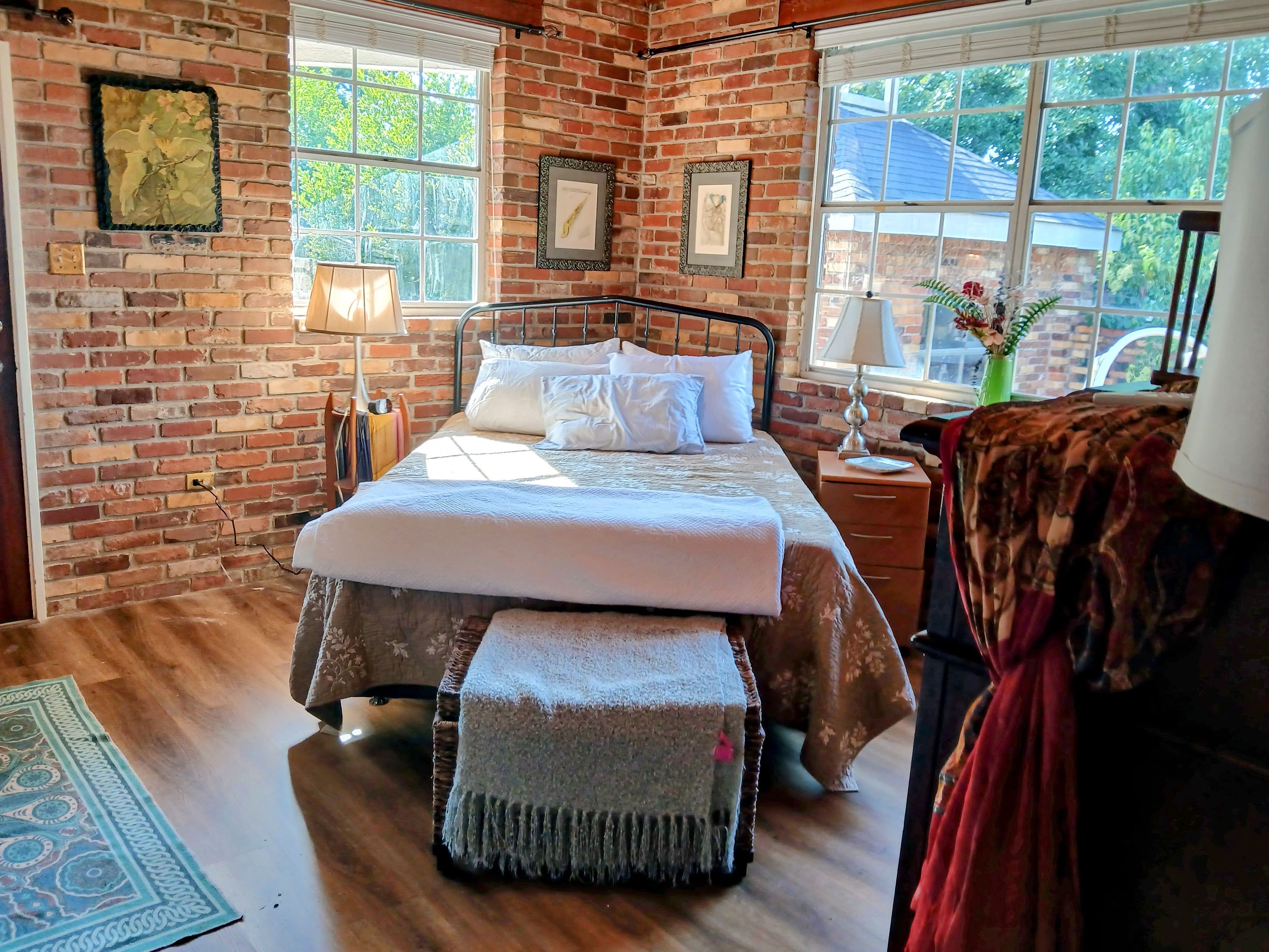A bedroom with a brick wall, a bed with white bedding, a bench, a nightstand with a lamp, and a window with a view of greenery.