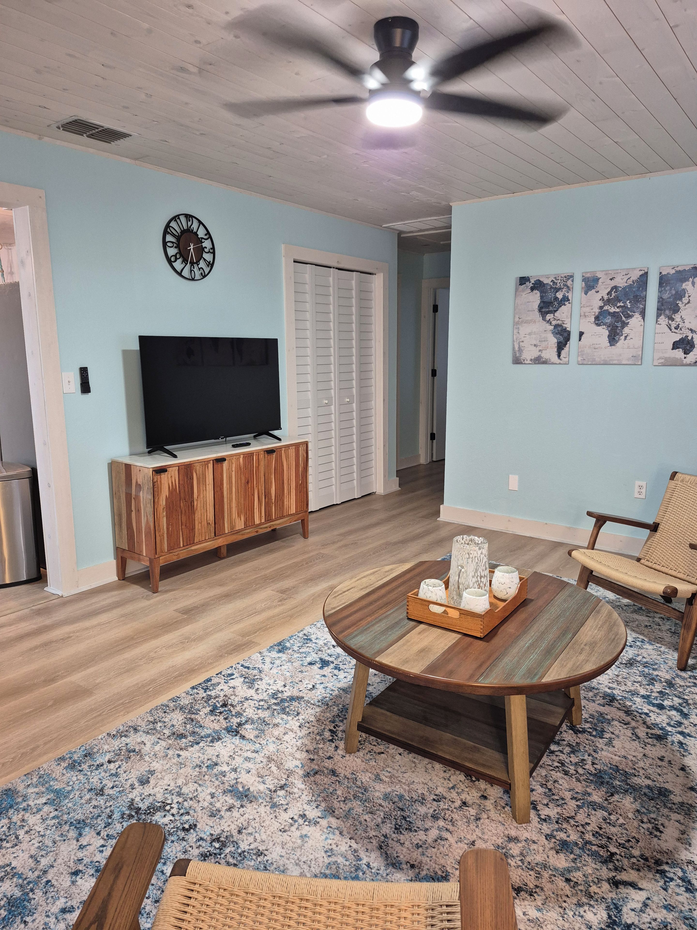 A living room with a wooden coffee table, a TV, a wooden cabinet, a woven chair, a world map artwork, and a ceiling fan.