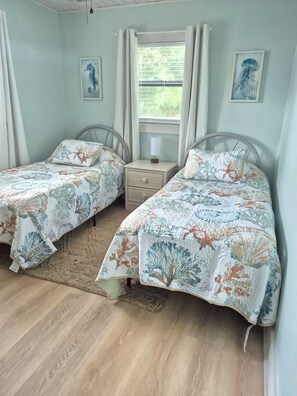 A bedroom with two single beds, a nightstand, a window with curtains, and wall art. - Almosta Beach House  in Clearwater (Clearwater)