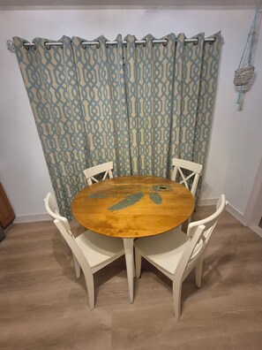 Une table ronde en bois, quatre chaises blanches, un rideau à motifs et un panier fixé au mur.