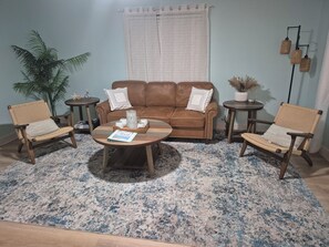 Un salon avec un canapé en cuir marron, deux fauteuils en osier, une table basse sur laquelle se trouve un livre, et un grand tapis aux motifs bleus et blancs.
