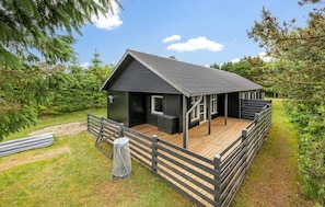 Une petite cabane en bois de couleur sombre, dotée d’une terrasse en bois et d’un espace clos, le tout entouré de verdure.