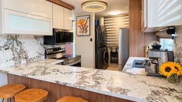 A modern kitchen with a marble countertop, stainless steel appliances, wooden bar stools, and a coffee machine.