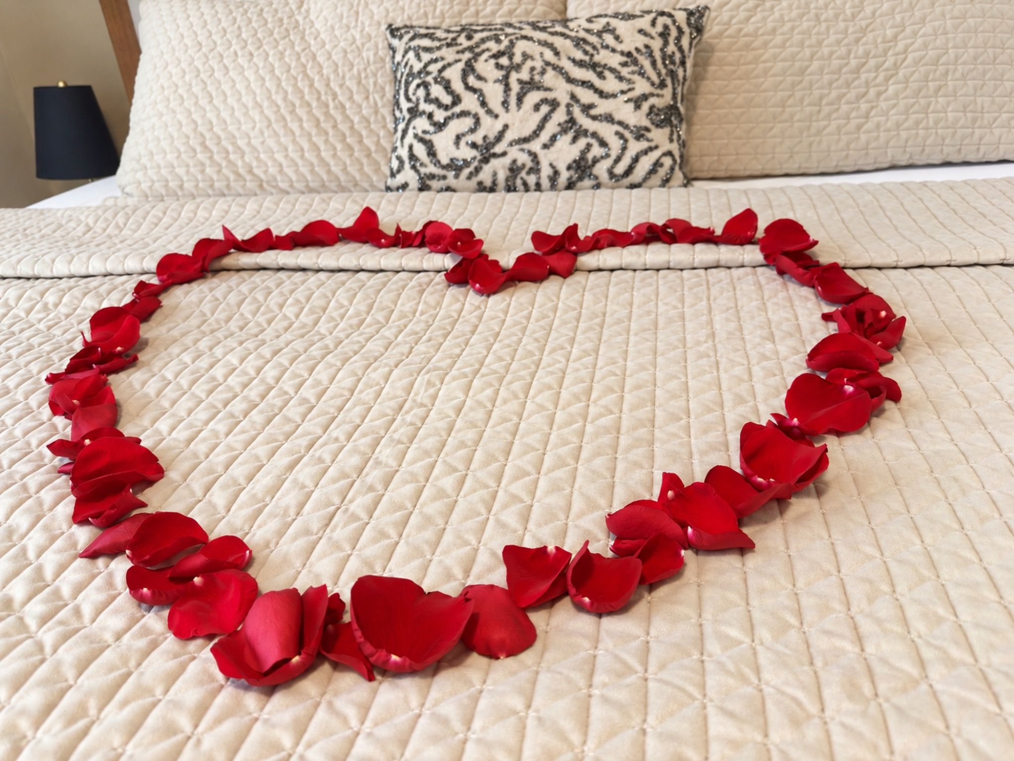 A bed with a quilted bedspread and a heart-shaped arrangement of red rose petals.