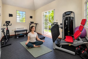 A gym with exercise equipment, a seated person meditating, and a view of greenery outside. - Ocean Views | 2 King Beds, Pool & Beach Gear (Rincón)