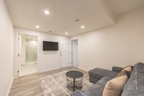 Ein modernes Wohnzimmer mit einem grauen Sofa, einem runden Couchtisch und einem flachbildfernseher, der an der Wand befestigt ist. Man sieht zwei weiße Türen und eine Duschkabine aus Glas.
