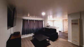 Ein modernes Wohnzimmer mit Fernseher, dunklen Vorhängen, einer Couch, einem Couchtisch und einer Lampe.