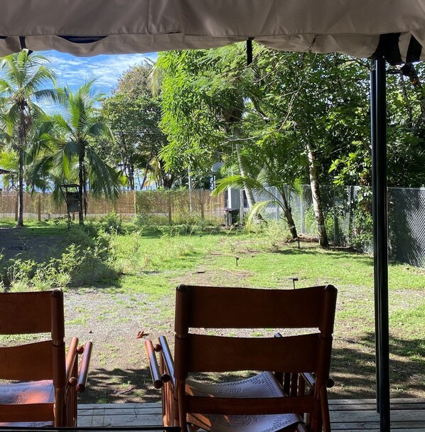 A covered outdoor seating area with wooden chairs, a view of a grassy yard with trees, and a fence in the background.