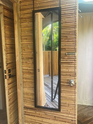 A room with a bamboo wall, a door with a mirror, and a view of a wooden deck and greenery outside.