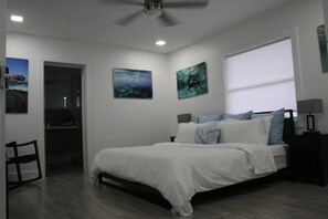 A bedroom with a bed, a desk, and a chair. There are paintings on the walls and a window with blinds.
