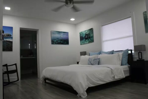 A bedroom with a bed, a desk, and a chair. There are paintings on the walls and a window with blinds.