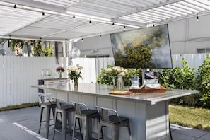 A covered outdoor kitchen area with a large island, bar stools, and a flat-screen TV.