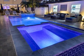 A swimming pool area with multiple pools, lounge chairs, and a seating area with sofas and a TV.