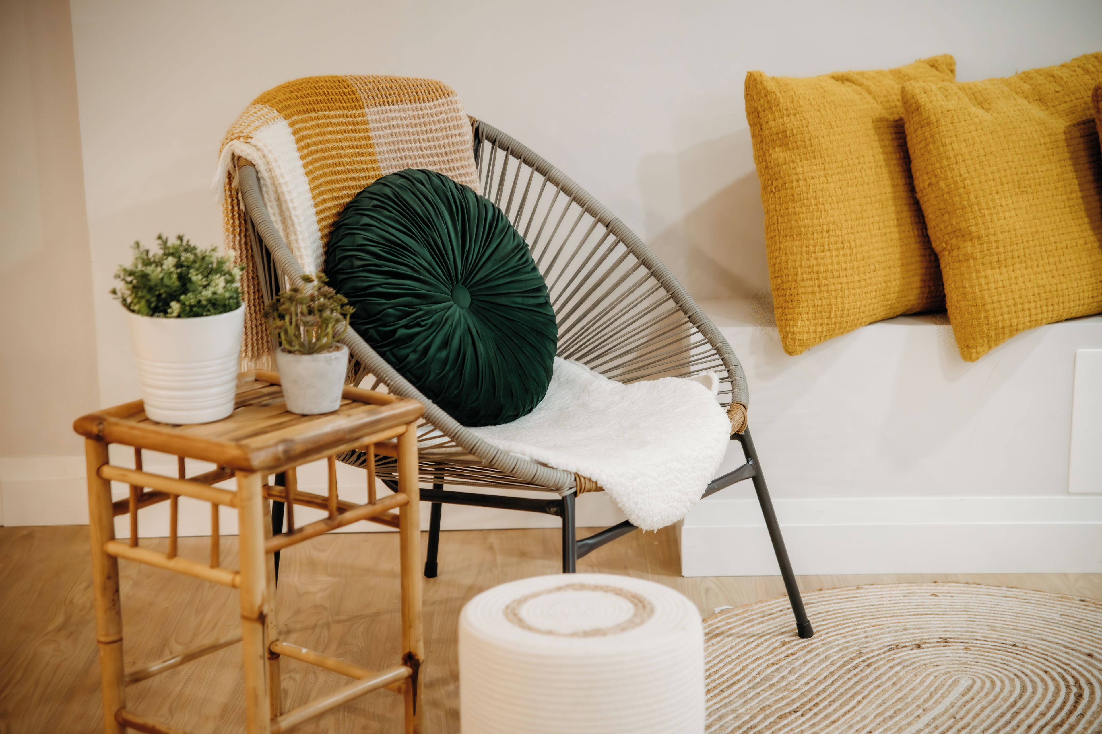 A wicker chair with a white cushion and a green pillow, a small wooden table with potted plants, and a woven rug on a wooden floor.