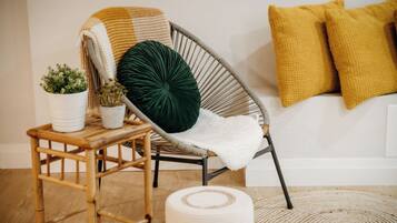 A wicker chair with a white cushion and a green pillow, a small wooden table with potted plants, and a woven rug on a wooden floor.