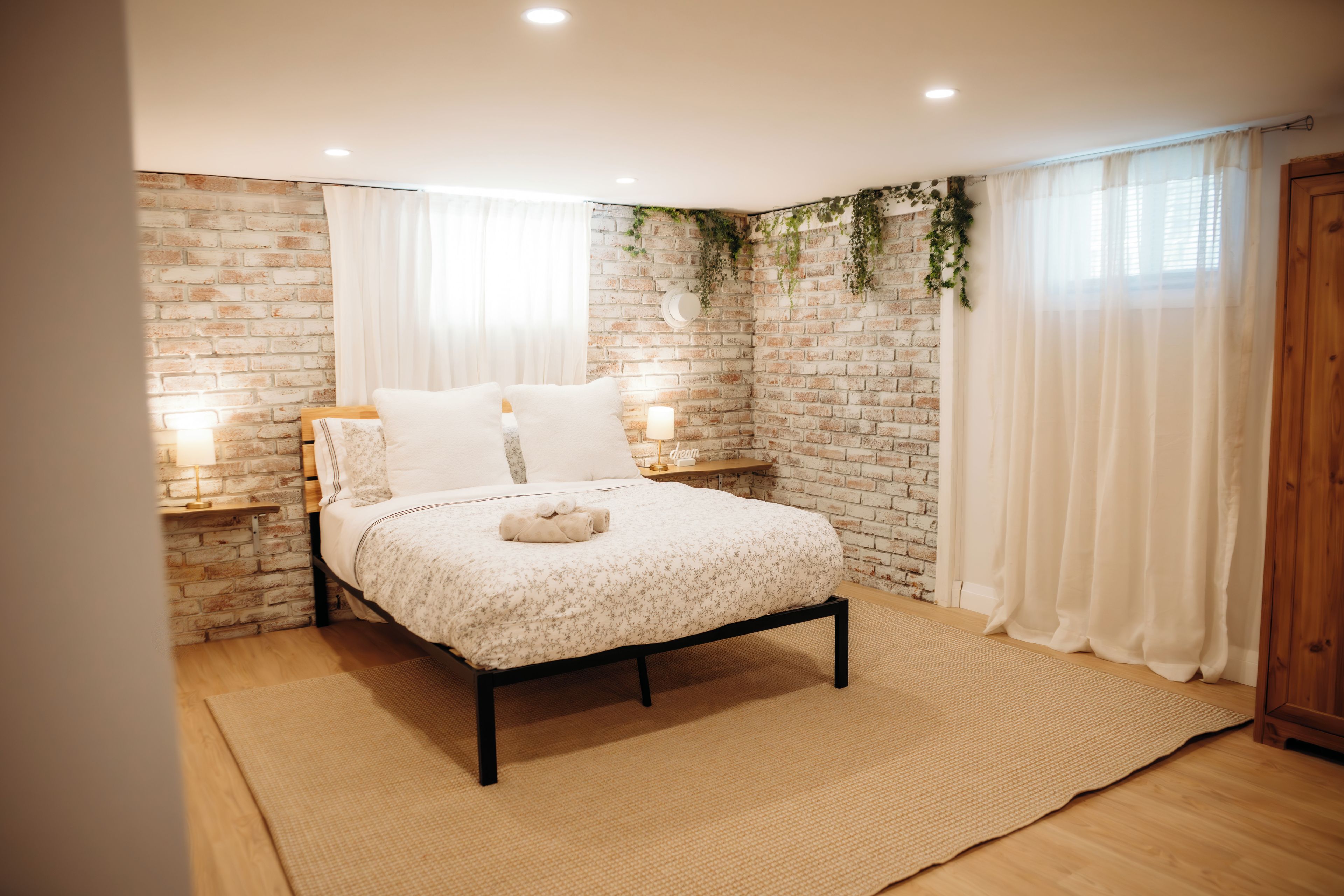 Une chambre avec un mur en briques, un lit recouvert d’une couvre-lit à motifs floraux, deux lampes de chevet et une fenêtre avec des rideaux.