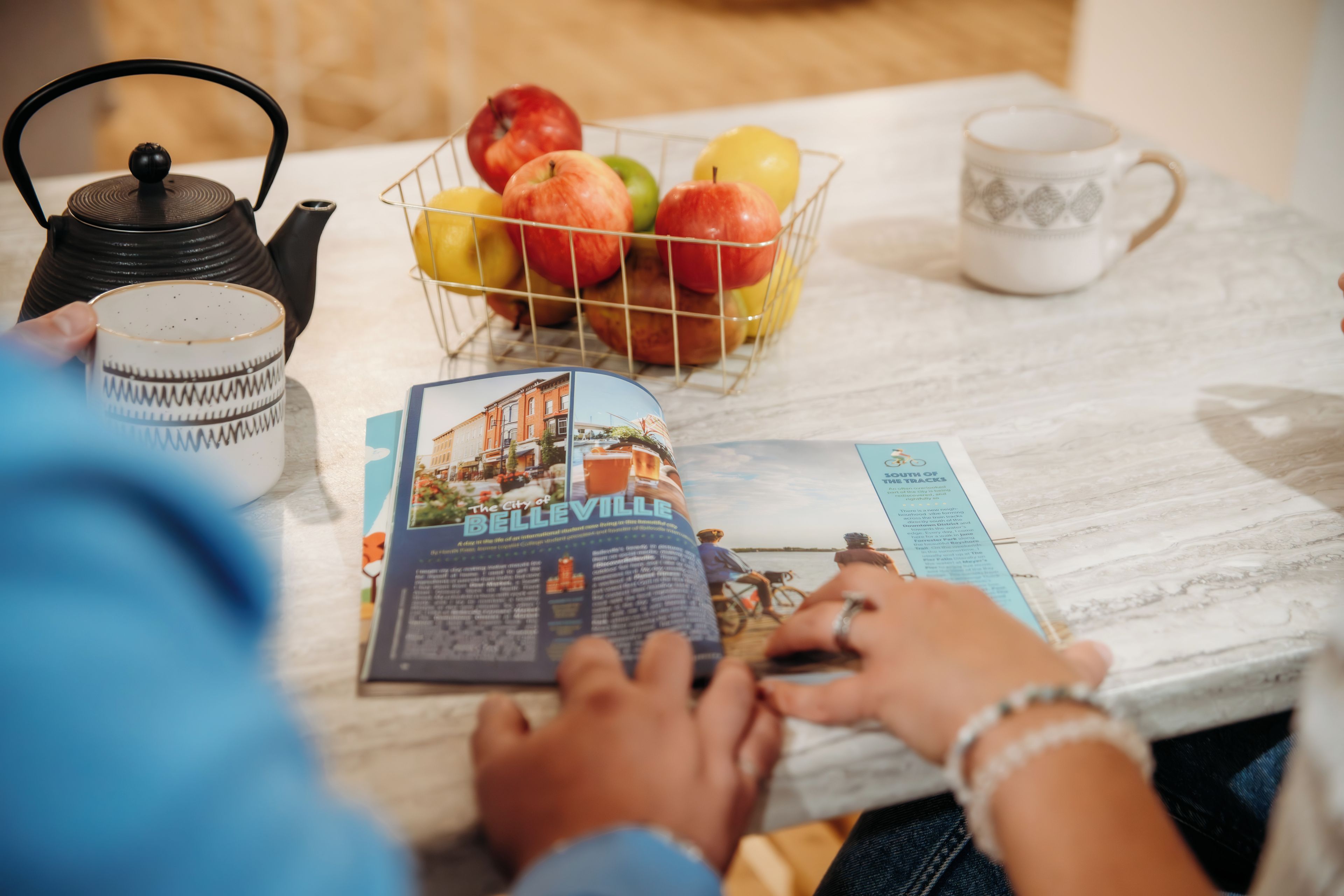 Una persona leyendo un libro junto a una mesa dispuesta con un set de té, una taza, una cesta de frutas y un plato decorativo.