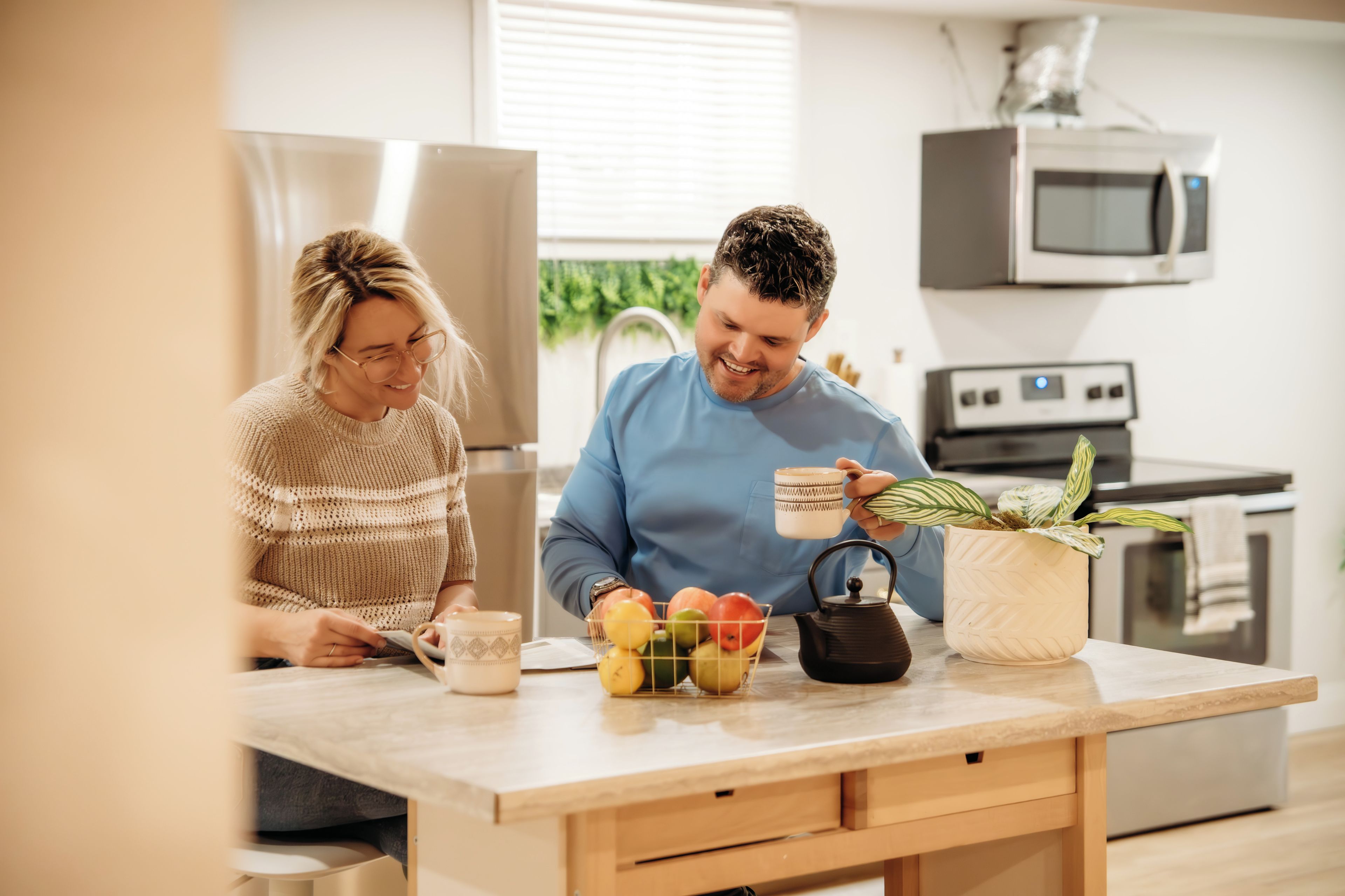 Une cuisine avec une table en bois, une bouilloire, une plante en pot et un panier de fruits.