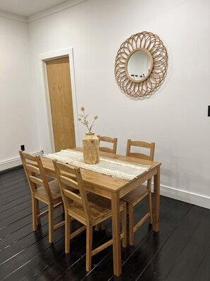 Un comedor con una mesa de madera, cuatro sillas, un espejo tejido y un jarrón con flores secas.