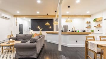 Una sala de estar moderna con un sofá gris, una mesa de comedor de madera y una zona de cocina con encimera de madera y lámparas colgantes.