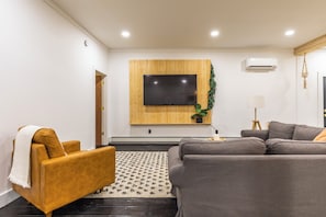 Una sala moderna con un televisor de pantalla plana grande montado en una pared de paneles de madera, un sofá gris, un sillón marrón y una alfombra estampada.