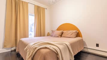 Une chambre avec un grand lit, des rideaux de couleur beige, un luminaire au plafond à motifs géométriques et une vue sur l’extérieur.