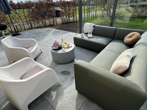 Ein moderner Außenbereich mit einer Sitzecke, einer Couch, zwei Stühlen und einem runden Tisch mit einer Flasche und Blumen.