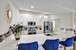 A modern kitchen with white cabinetry, stainless steel appliances, and a central island with bar stools. - Tropical Beachside Bliss – Pool, Tiki Hut & Bar (Fort Lauderdale)