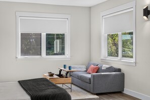 A modern living room with a grey sofa, a wooden coffee table, and a black ottoman. There are two windows with white blinds, and a small table with a blue mug and a brown bowl. - Sugar Bridge Loft · Studio Steps from Trails (Bella Vista)