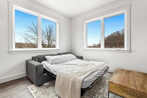 A bedroom with a bed, a gray sofa, a wooden table, and two windows with a view of trees and a building. - Silent Hattie Loft · Studio by Tunnel Vision Trail (Bella Vista)