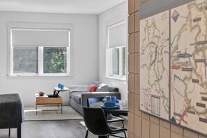 Un salon moderne avec un canapé, une table basse et un coin repas avec des chaises. Il y a des fenêtres qui laissent entrer la lumière naturelle et une carte est accrochée au mur.