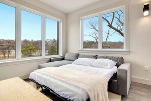 A bedroom with a bed, a desk, and large windows offering a view of trees and a clear sky. - Play Thru Loft · 2BR w/ Balcony Near Back 40 (Bella Vista)