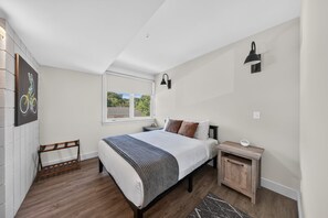 A bedroom with a bed, a nightstand, a wall-mounted lamp, a window, and a bicycle-themed artwork. - Staggerwing Loft · 2BR Above Bike Shop & Café (Bella Vista)