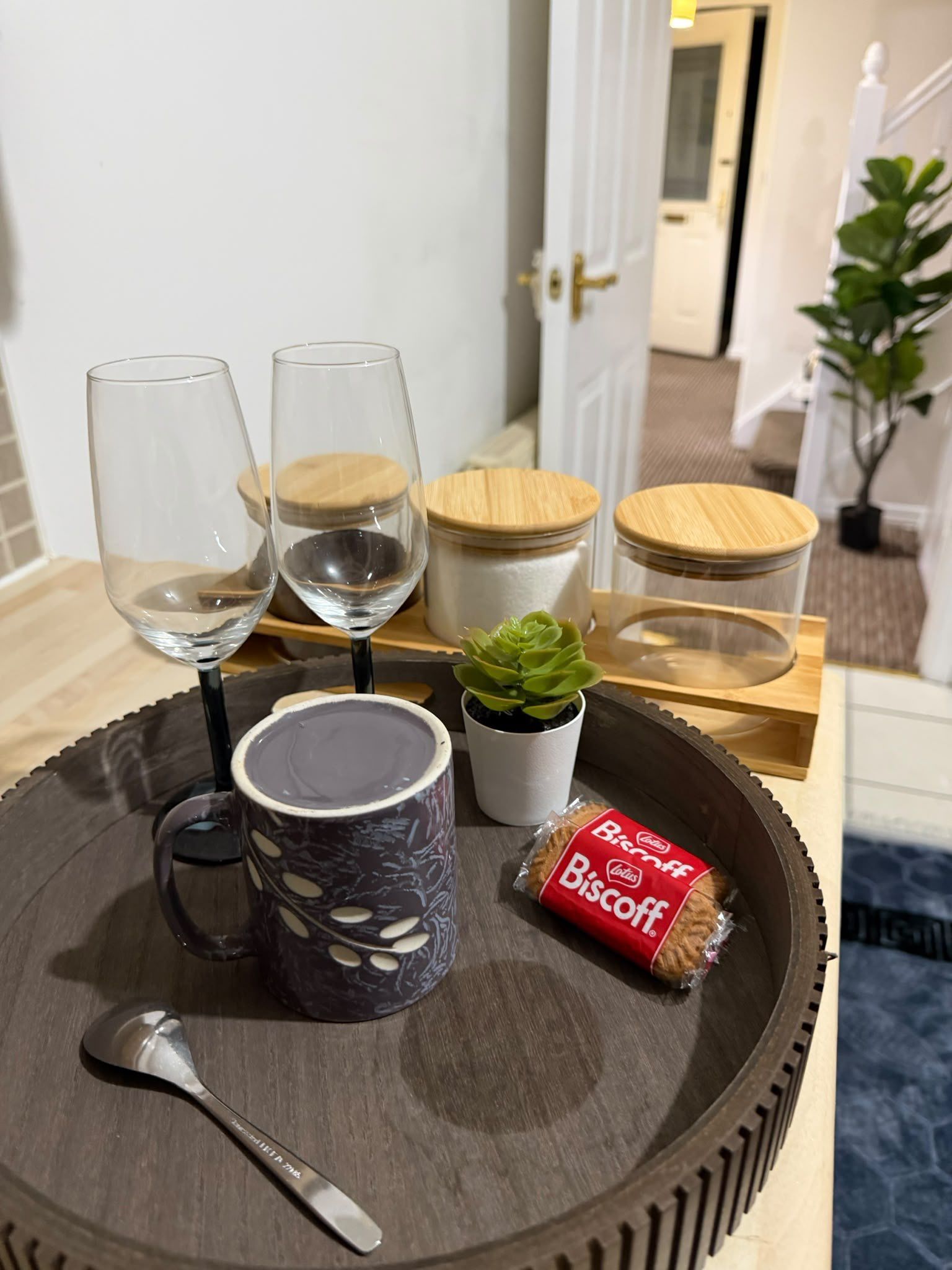A tray with a glass, a cup, a biscuit, a spoon, and a small plant on a wooden surface.