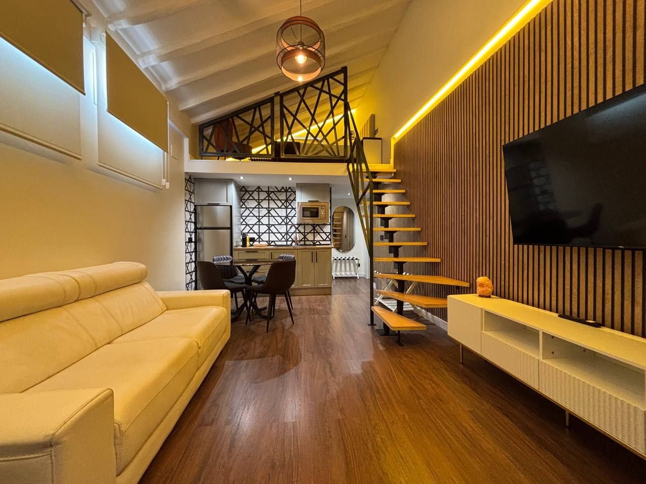 A modern living room with a sofa, dining table, chairs, a TV, and a staircase leading to an upper level.