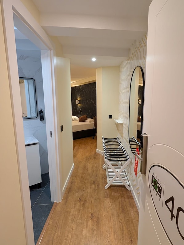 A hotel corridor with a wooden floor, a bed with a dark headboard, a mirror, and a row of striped chairs.