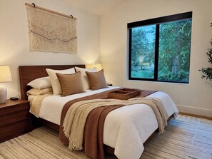 Quarto com uma cama grande, mesa de cabeceira, painel de macramé fixado na parede e janela com vista para árvores.