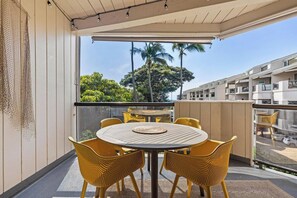 Ein Balkon mit einem runden Tisch und gelben Korbstühlen, der auf einen Wohnbereich mit Palmen und einem klaren Himmel blickt.
