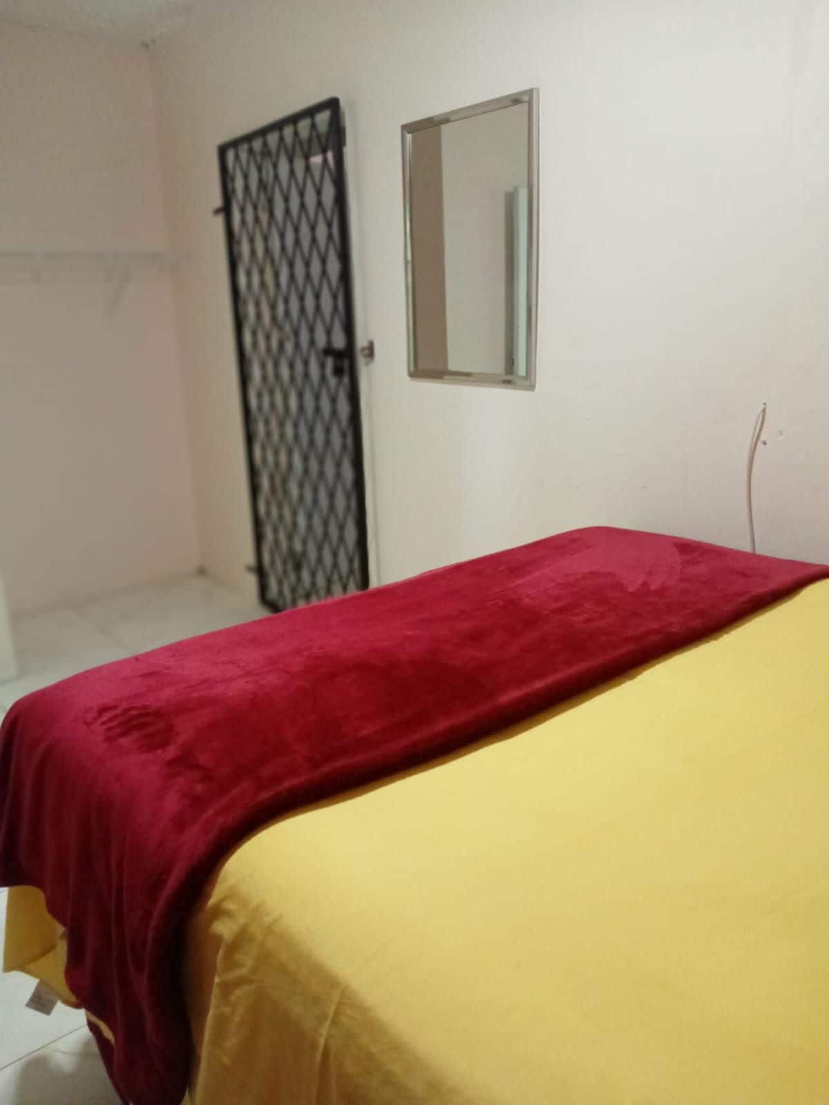Ein Zimmer mit einem Bett, das mit einer roten und gelben Decke bezogen ist, einer verschlossenen Tür mit einem Metallgitter und einem kleinen Spiegel an der Wand.