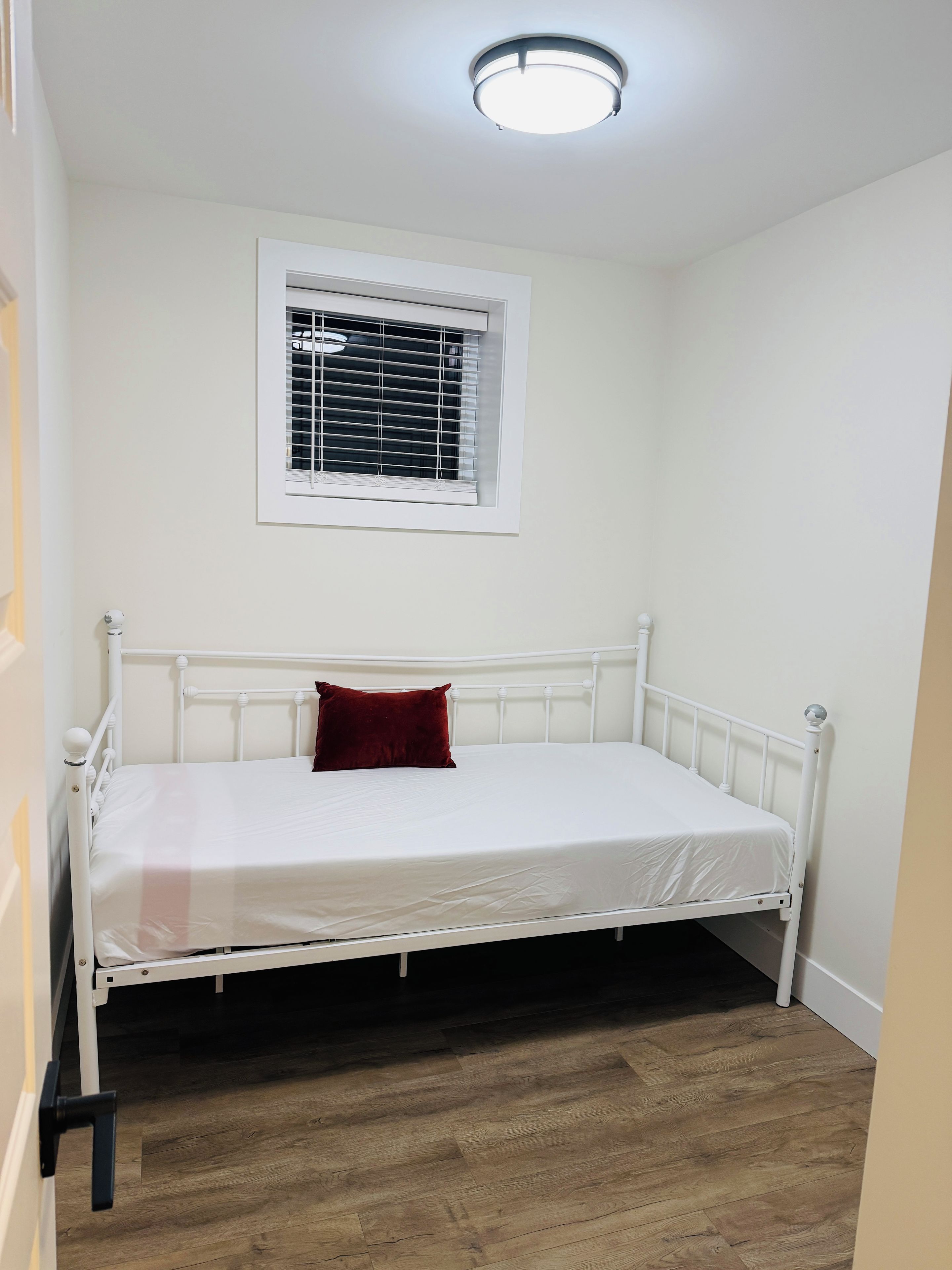 A single bed with a white frame and a red pillow, a window with blinds, and a wooden floor.