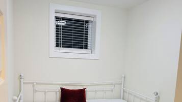 A single bed with a white frame and a red pillow, a window with blinds, and a wooden floor.