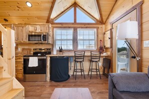 A wooden cabin interior with a kitchen, two bar stools, a sofa, and a large window. - Firelight Cabin | Loft + 2 Baths | Sleeps 8 (Fredericksburg)